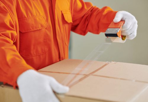 Unrecognizable house moving service worker wearing bright orange uniform closing box using tape